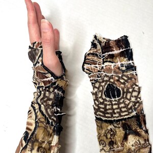May include: A pair of fingerless arm warmers featuring a layered, ruffled construction. The fabric displays a repeating abstract pattern in shades of brown, black, and cream. Each sleeve has a thumb hole and a textured, tiered design.