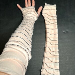 May include: A pair of pale pink arm warmers featuring a layered, ruffled design with thin, dark grey lettuce-edge trim along each horizontal tier. The fabric is semi-sheer and lightweight, extending from the fingers up to the upper arm area.
