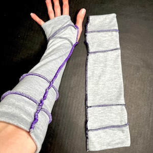 May include: Gray arm warmers with purple stitching. One warmer is on a hand, the other is separate. The warmers are made of a soft material and feature decorative stitching along the seams and edges.