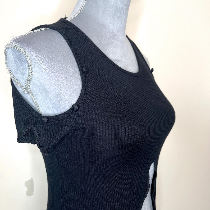May include: Black ribbed tank top with a scoop neckline and shoulder cutouts. The tank top has small buttons on the shoulders and a slit up the side. The fabric has vertical ribbing.