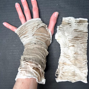 May include: A pair of distressed, fingerless gloves. The gloves are made of a light beige fabric with a shredded design. The fabric is torn in multiple places, creating a tattered appearance. The gloves are designed to cover the wrist and part of the hand.