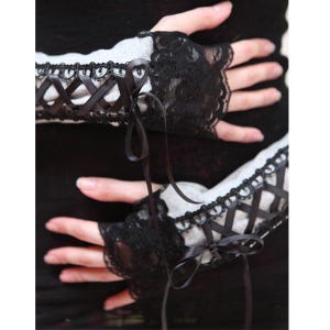 May include: A pair of fingerless gloves featuring a light gray fabric base with black lace trim at the edges. The gloves are decorated with a black ribbon corset-style lacing detail that runs along the forearm and ties into a bow at the wrist.