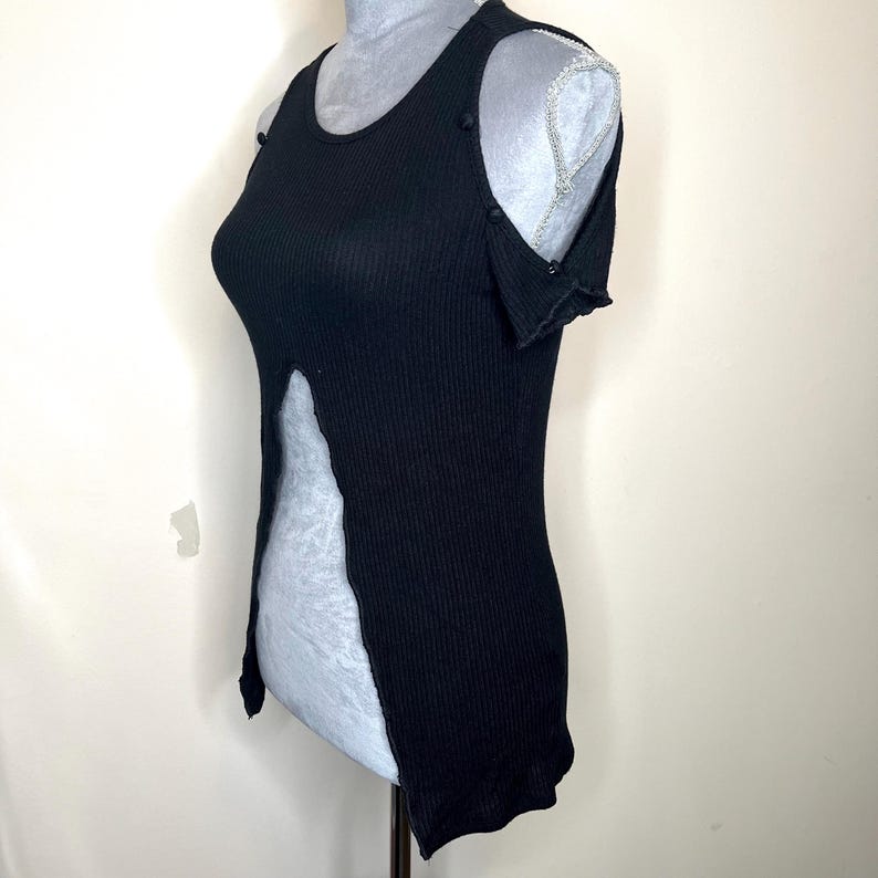 May include: Black ribbed tank top with a split front design. The sleeveless top features button details on the shoulders and a scoop neckline. The fabric has a vertical ribbed texture.