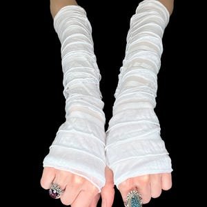 May include: A pair of long, white, sheer arm warmers featuring a gathered, ruched texture that extends from the wrist to the upper forearm. The fabric is lightweight and semi-transparent, creating horizontal folds along the entire length of the sleeves.
