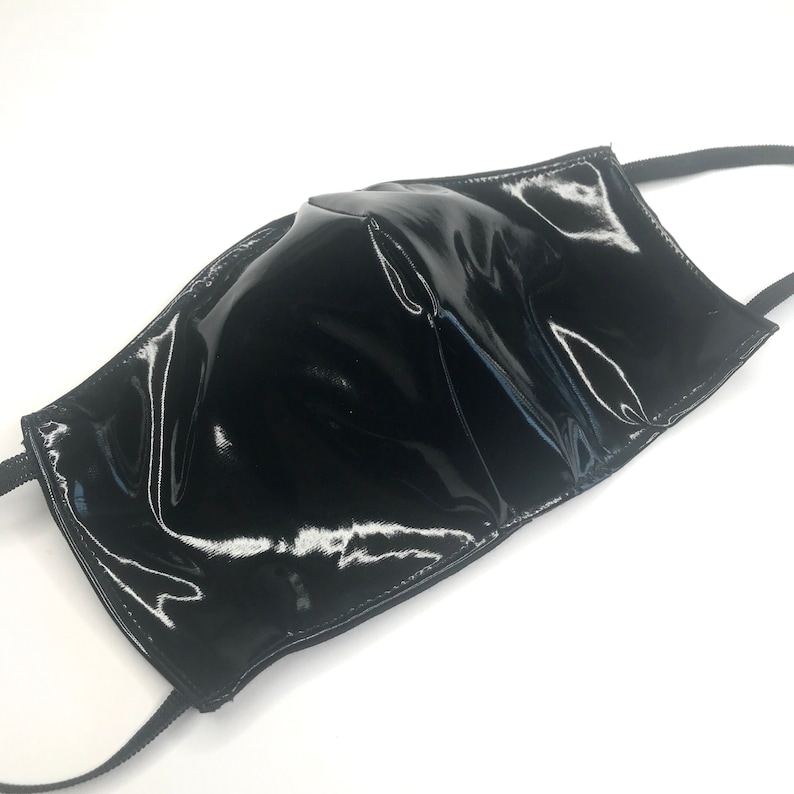 May include: A black face mask with a high-gloss, patent-like finish. The mask features a structured, contoured center seam and is attached to thin, black elastic ear loops on both sides, providing a sleek and reflective appearance throughout.