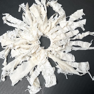 Könnte beinhalten: Ein handgefertigter, cremefarbener Stoffkranz mit kreisförmiger Mitte und strahlenförmigen Streifen unterschiedlicher Texturen. Die Streifen umfassen Spitze und ausgefranste Kanten, wodurch eine strukturierte, böhmische Ästhetik entsteht. Der Kranz ist vor einem dunklen Hintergrund platziert.