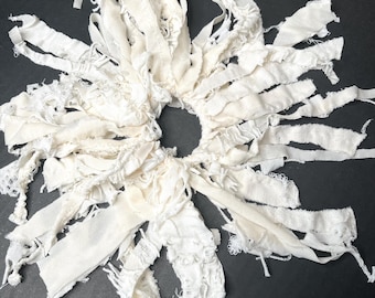 May include: A handmade, off-white fabric wreath with a circular center and radiating strips of varying textures. The strips include lace and frayed edges, creating a textured, bohemian aesthetic. The wreath is set against a dark background.