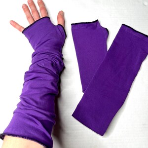 May include: A pair of vibrant purple fingerless gloves that extend up the arm. The gloves feature a solid purple fabric construction with a simple thumb hole opening and a thin black stitched trim along the edges of the cuffs and thumb hole.