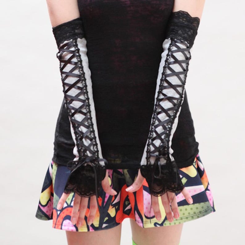 May include: A pair of light gray arm warmers featuring black lace trim at the top and bottom edges, with a black ribbon crisscross lacing design running down the center of each sleeve, finished with small black ribbon bows at the wrists.