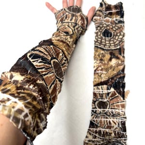 May include: A pair of long, fingerless arm sleeves featuring a textured, crinkled fabric with a brown, black, and cream abstract circular pattern. The sleeves extend from the fingers up the arm, showcasing a repeating geometric design.