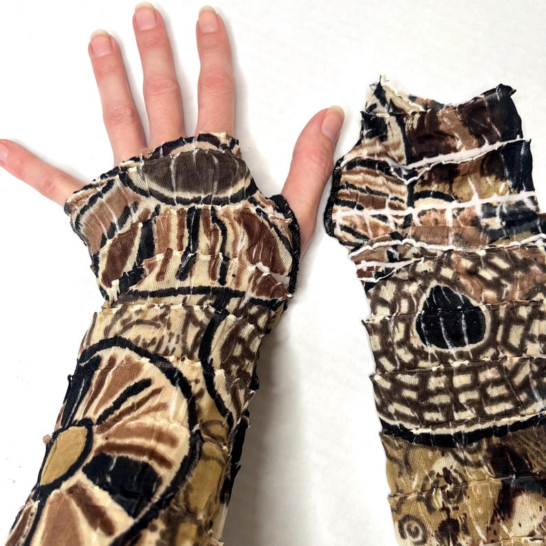 May include: A pair of fingerless arm warmers featuring a crinkled, textured fabric with a repeating geometric and circular pattern in shades of brown, black, and cream. The sleeves have a layered, ruffled appearance with raw, unfinished edges.