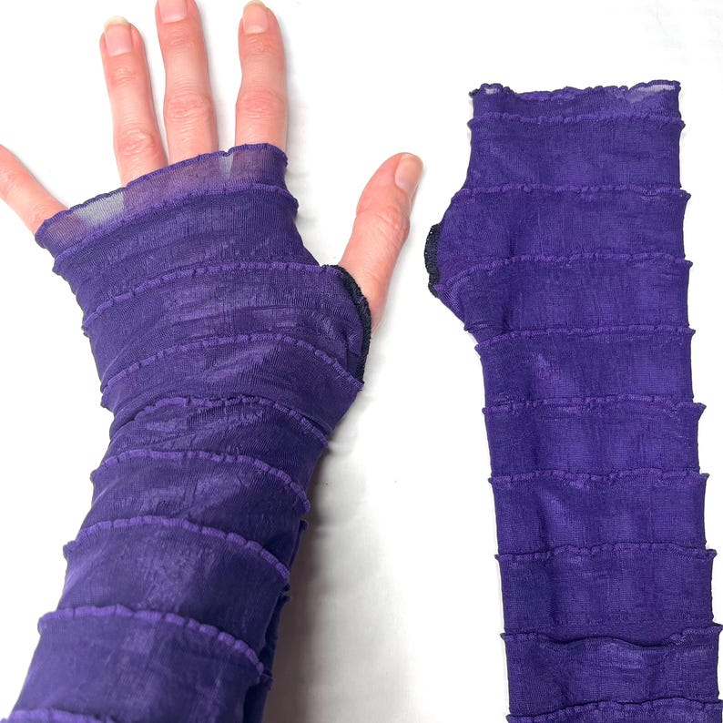 May include: A pair of fingerless arm warmers in a deep purple hue, crafted from a sheer, crinkled fabric. The design features horizontal ruffled layers throughout the length of the sleeves, with a dedicated opening for the thumb on each piece.
