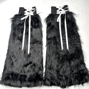 May include: A pair of black faux fur leg warmers featuring a white ribbon lace-up detail at the top. The long, shaggy black synthetic fur covers the length of the leg warmers, while the white satin ribbons are threaded through eyelets and tied.