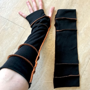 May include: A pair of black arm warmers with orange stitching. The arm warmers are made of a soft, stretchy fabric and have a ribbed texture.