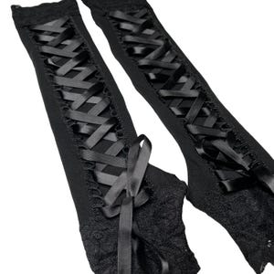 May include: A pair of black fingerless gloves featuring a long, vertical lace-up design with satin ribbon threaded through eyelets. The gloves are finished with intricate black lace trim at the wrist and a decorative ribbon bow at the base.