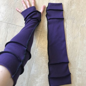 May include: A pair of purple arm warmers with black stitching. The arm warmers are made of a soft, stretchy fabric and have a fitted design.