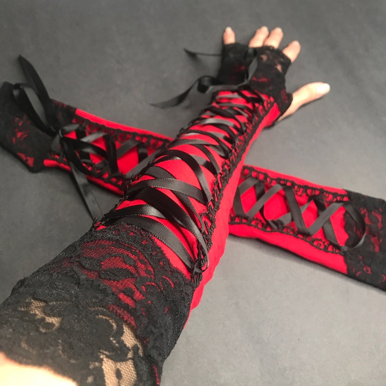 May include: A pair of fingerless arm warmers in vibrant red fabric, featuring black lace trim at the edges and a decorative black ribbon corset-style lacing system that runs along the length of the forearm, secured with matching black ribbon ties.