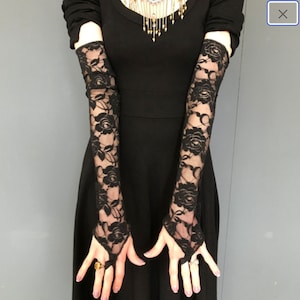 May include: A pair of black lace fingerless gloves with floral designs. The gloves are worn by a person wearing a black dress.