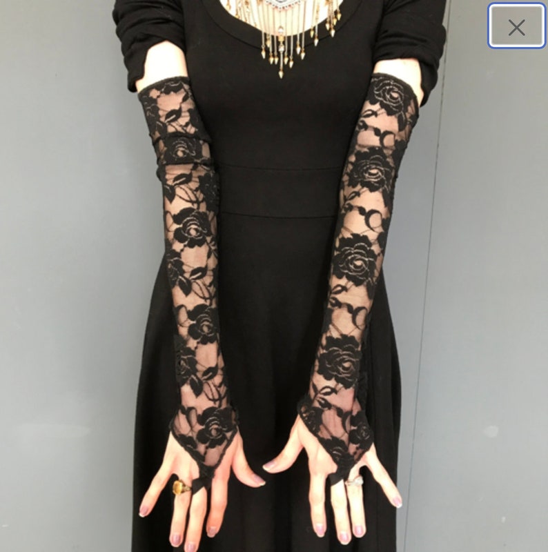 May include: A pair of black lace fingerless gloves with floral designs. The gloves are worn by a person wearing a black dress.