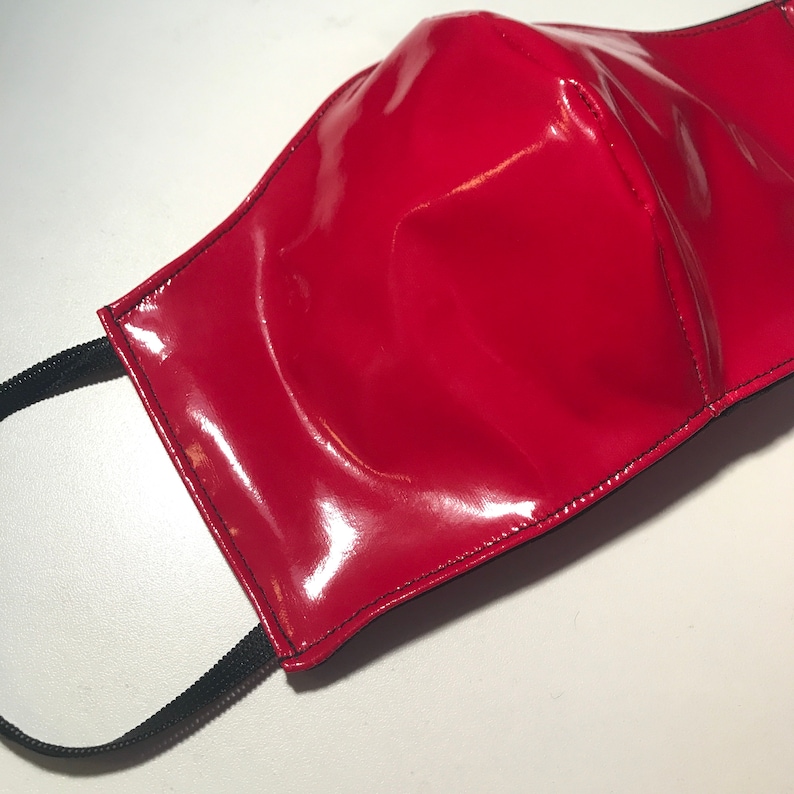 May include: A bright red face mask with a glossy, patent-like finish. The mask features a central vertical seam and is constructed with black stitching along the edges. It includes black elastic ear loops attached to each side of the mask.