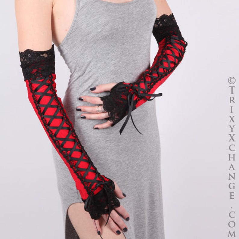May include: Red and black arm warmers with a gothic aesthetic. The arm warmers feature a red fabric base, criss-cross black ribbon detailing, and black lace trim at the top and around the fingerless hand openings. The design includes black bows.