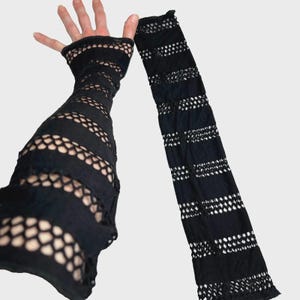 May include: Black, open-weave arm warmers with a fingerless glove design. The fabric features horizontal stripes and a dotted pattern, creating a textured look. The arm warmers are made of a stretchy material.