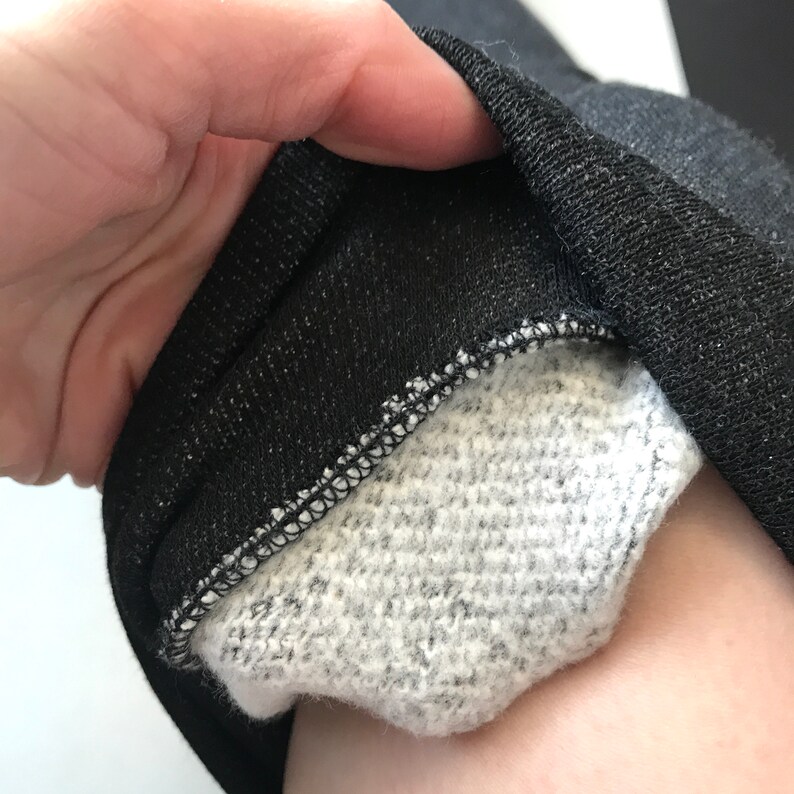 Puede incluir: Primer plano de un pu&ntilde;o de sudadera negro con un interior gris y afelpado. El pu&ntilde;o est&aacute; doblado, revelando el forro suave y texturizado y las costuras a lo largo del borde. La tela tiene un brillo sutil.