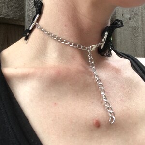 May include: A black lace choker with a silver chain and a silver clasp. The choker is worn around a person's neck.
