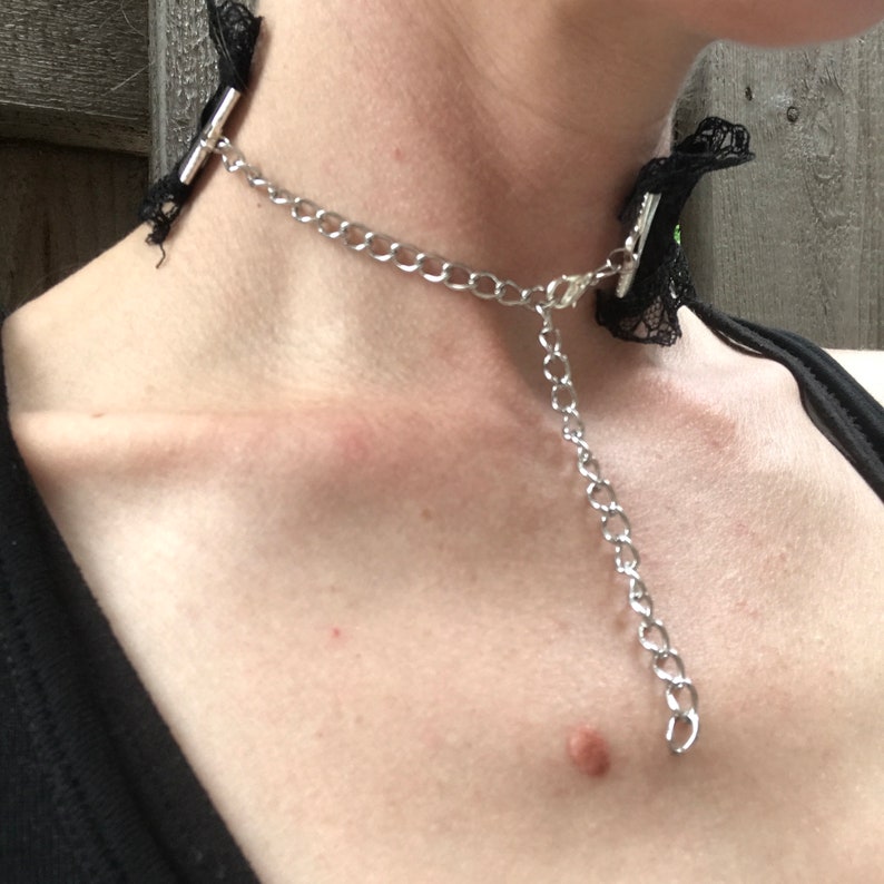 May include: A black lace choker with a silver chain and a silver clasp. The choker is worn around a person's neck.