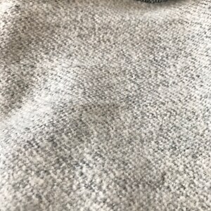 Puede incluir: Primer plano de una tela texturizada gris y blanca. El material tiene un aspecto moteado con un patr&oacute;n tejido. La tela parece suave y posiblemente hecha de lana o un material similar.