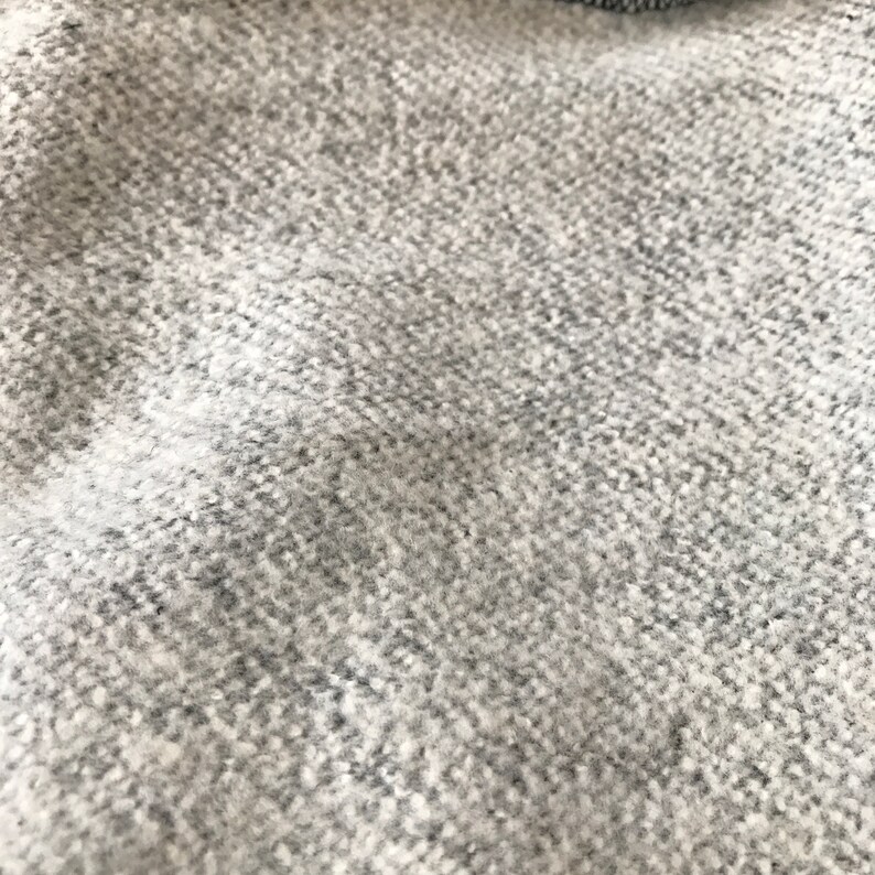Puede incluir: Primer plano de una tela texturizada gris y blanca. El material tiene un aspecto moteado con un patr&oacute;n tejido. La tela parece suave y posiblemente hecha de lana o un material similar.