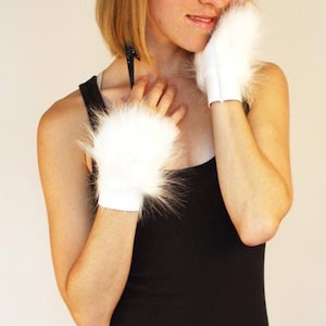 May include: A pair of white fingerless gloves featuring a plush, long-pile faux fur texture on the back of the hands and a smooth, solid white fabric cuff at the wrist. The gloves are designed to cover the hand while leaving the fingers exposed.