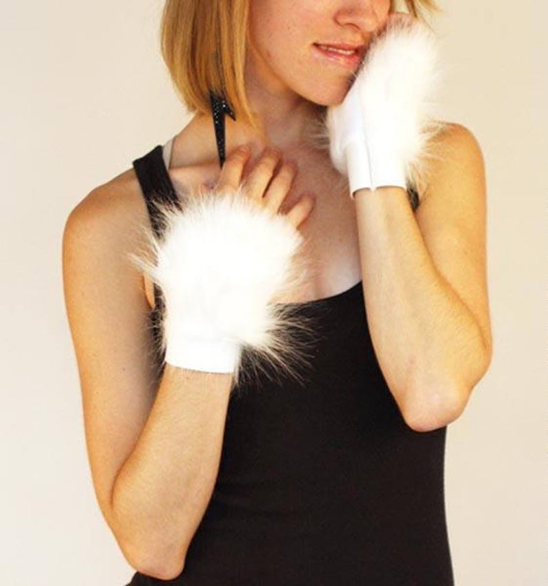 May include: A pair of white fingerless gloves featuring a plush, long-pile faux fur texture on the back of the hands and a smooth, solid white fabric cuff at the wrist. The gloves are designed to cover the hand while leaving the fingers exposed.