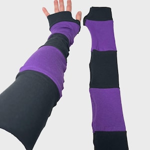May include: A pair of arm warmers featuring alternating horizontal stripes of black and purple fabric. The arm warmers are designed with a thumb hole opening at the top, extending down the arm to provide coverage from the wrist to the upper forearm.