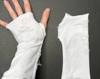 May include: A pair of white cotton fingerless gloves with distressed detailing. The gloves are shown laid flat and on a hand.