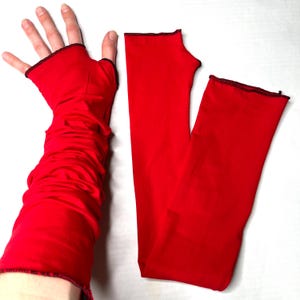 May include: A pair of bright red arm warmers featuring a fingerless design with a thumb hole. The fabric is smooth and flexible, with a subtle gathered texture on the arm. Each opening is finished with a thin, dark, contrasting stitched edge.