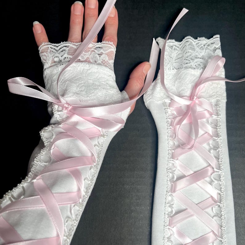 May include: A pair of white fingerless gloves featuring a lace trim at the top and a corset-style lace-up design down the center. The gloves are adorned with a light pink satin ribbon woven through white eyelet loops in a crisscross pattern.