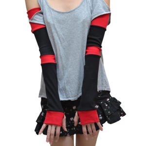 May include: A grey scoop-neck top with cold-shoulder cutouts, paired with long arm warmers featuring alternating horizontal bands of black and red fabric. The outfit includes a black mini skirt with a small floral pattern and tiered ruffled hem details.