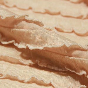 May include: A light peach-colored fabric featuring horizontal rows of gathered ruffles with a lettuce-edge finish. The material has a fine, ribbed texture and is arranged in soft, overlapping folds that create a layered, wavy appearance throughout.