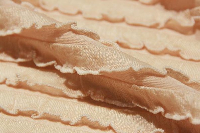 May include: A light peach-colored fabric featuring horizontal rows of gathered ruffles with a lettuce-edge finish. The material has a fine, ribbed texture and is arranged in soft, overlapping folds that create a layered, wavy appearance throughout.