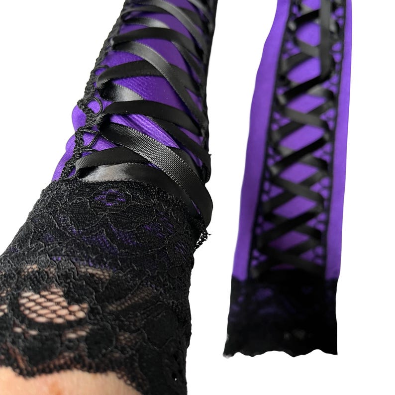 May include: Purple and black arm warmers with a lace cuff. The arm warmers feature a purple fabric base, crisscrossed with black satin ribbons. The cuffs are made of black lace.