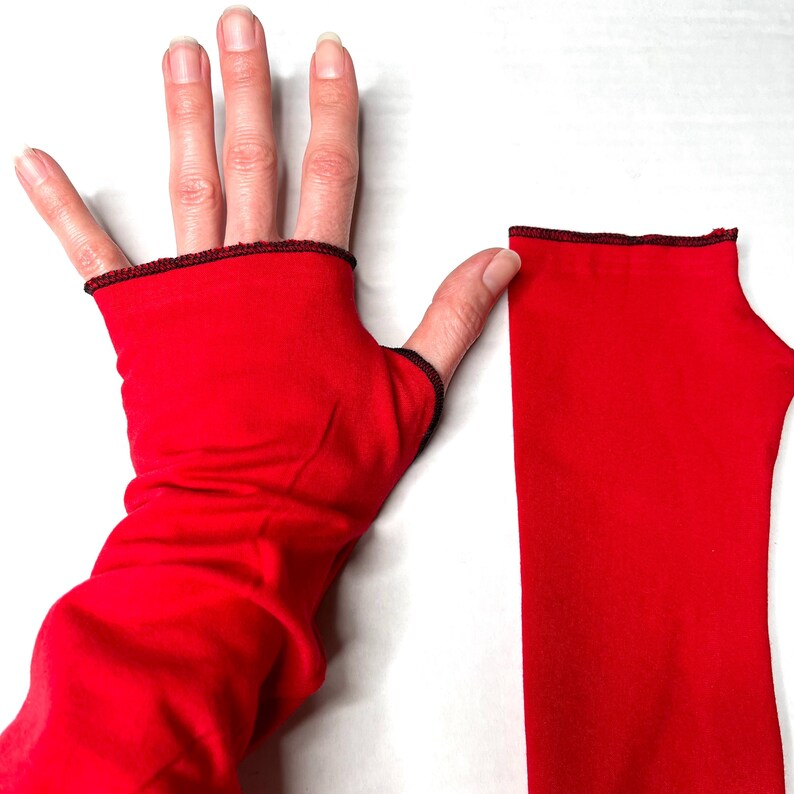 May include: A pair of bright red fingerless gloves made of a smooth, stretchy fabric. Each glove features a simple thumb hole and is finished with a thin, dark maroon or black serged edge along the top opening and the thumb opening for a clean look.