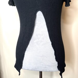 May include: A black, ribbed knit top with a unique open back design. The top features short, ruffled sleeves and a split that extends down the back, creating a flowing silhouette. The hemline has a scalloped edge.