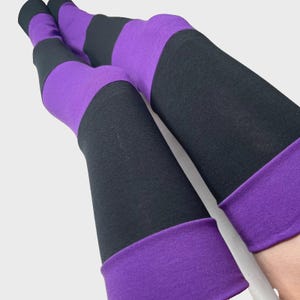 May include: A pair of over-the-knee socks featuring wide horizontal stripes in alternating solid black and vibrant purple colors. The fabric has a smooth, matte texture, and the socks are finished with a wide purple cuff at the top edge.