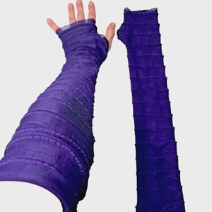 May include: A pair of long, deep purple fingerless gloves featuring a textured, ruched, or gathered fabric design that extends from the knuckles up the forearm. The material appears semi-sheer and lightweight, with horizontal ridges throughout.