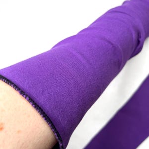 May include: A long, solid purple fabric sleeve featuring a finished hem with visible dark purple stitching. The material appears to be a soft, flexible textile that extends along the arm, showcasing a consistent deep violet color throughout.