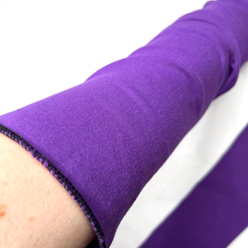 May include: A long, solid purple fabric sleeve featuring a finished hem with visible dark purple stitching. The material appears to be a soft, flexible textile that extends along the arm, showcasing a consistent deep violet color throughout.