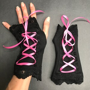 May include: A pair of black fingerless gloves with pink ribbon lacing. The gloves have a lace trim and are made of a soft, stretchy fabric.
