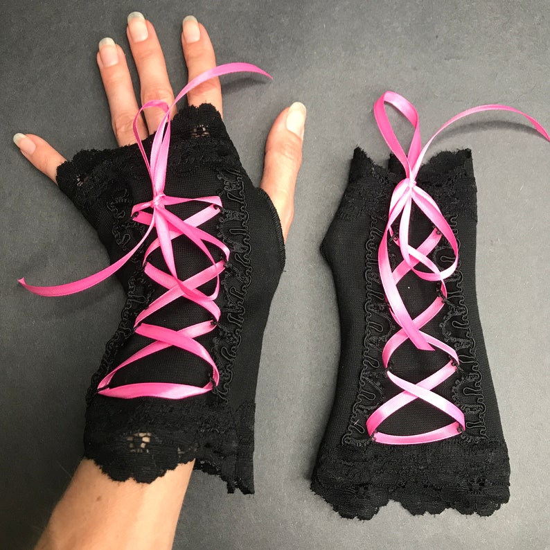 May include: A pair of black fingerless gloves with pink ribbon lacing. The gloves have a lace trim and are made of a soft, stretchy fabric.