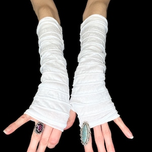Puede incluir: Un par de guantes blancos, transparentes y largos, que presentan una textura horizontal fruncida o recogida en toda la tela. Los guantes no tienen dedos, se extienden desde los nudillos hasta la mitad del antebrazo, con un material ligero y semitransparente.